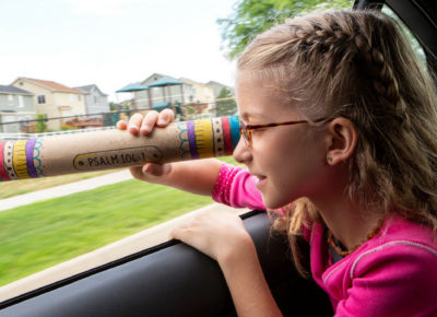 Elementary age girl is looking through a decorate paper towel tube out the car window.