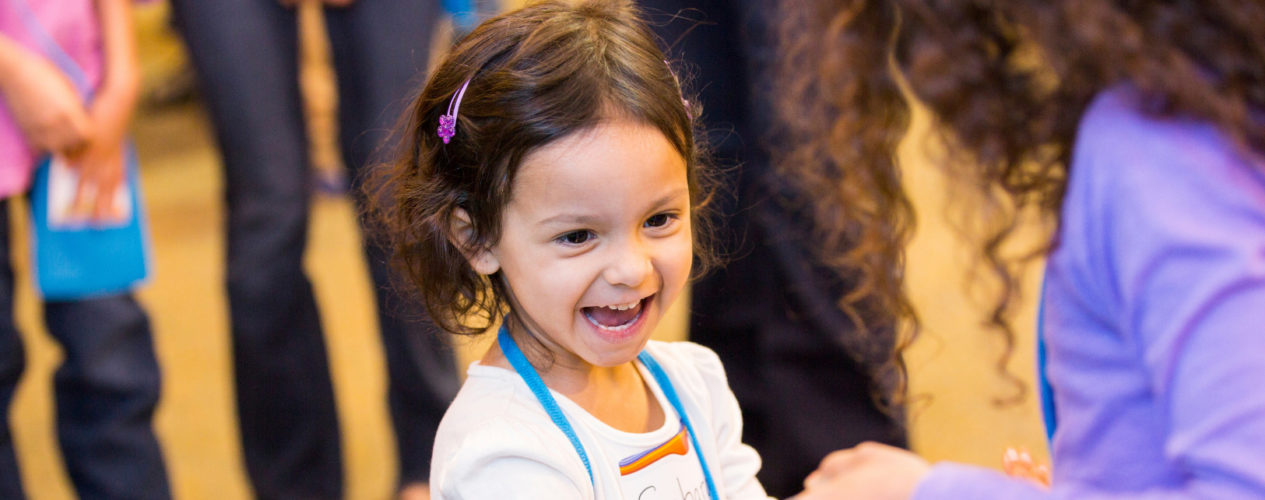 A preschool girl smiles joyfully as an older girl hands her something.