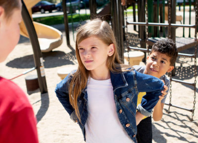 Girl in jean jacket standing with her hands on her hips as a boy stands scared behind her. She's standing up to a bigger boy facing the two of them.