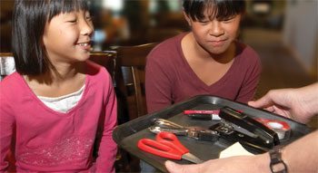Two children observe a tray with random objects on it.