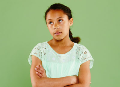 A preteen girl standing in front of a green wall. Her arms are crossed in front of her body and she's rolling her eyes.