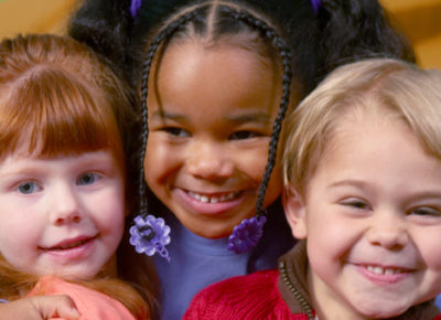 Three early elementary kids smiling as they hug one another in their classroom.
