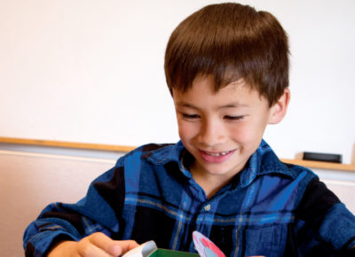 An elementary-aged boy is sitting at a table in his classroom participating in a bottle bug craft.