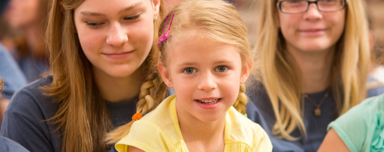 Little girl with blonde hair and pigtails stares directly at the camera and has a smirk on her face. She is sitting on a volunteers lap.