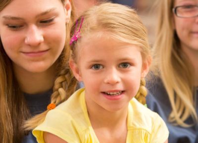 Little girl with blonde hair and pigtails stares directly at the camera and has a smirk on her face. She is sitting on a volunteers lap.