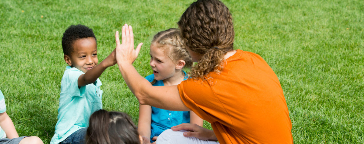 Adult leader high fives a preschool boy as she sits in a circle on the grass with her preschool class.