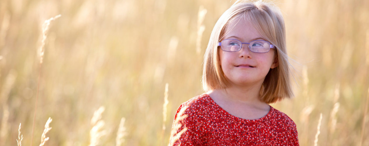 An elementary-aged girl with Down syndrome standing in a field smiling.