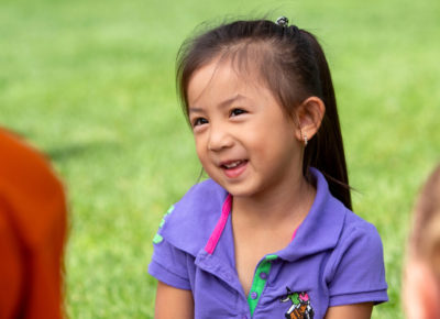 A preschool girl in a purple shirt is sitting on the grass among other preschoolers as they learn about a springtime craft they'll be doing.