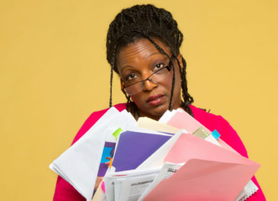 An exhuasted looking women holding a lot of disheveled folders. She's probably so exhausted because she didn't choose a published curriculum.