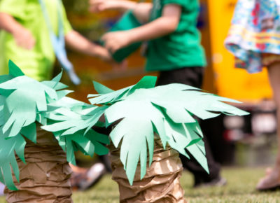 Two tiny palm trees constructed out of paper sit outside in the foreground as preschoolers run around in the background.