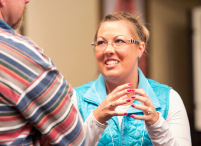 A woman in a bright blue vest talking to a man who's back is to the camera. She is talking with her hands as she smiles genuinely as she talks.