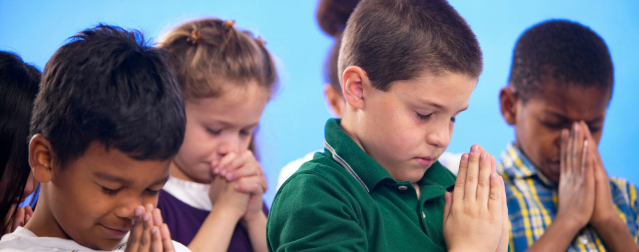 A group of children are praying with their eyes closed, heads bowed, and hands folded. They are standing in front of a blue wall.