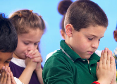 A group of children are praying with their eyes closed, heads bowed, and hands folded. They are standing in front of a blue wall.