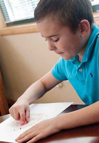 Elementary-aged boy erasing a page that is colored on with brown crayon.