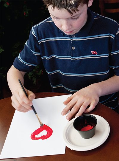 Preteen boy painting a red heart on a sheet of paper.