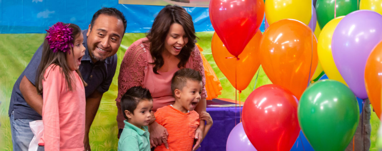 A family with a mom, dad, an elementary-aged daughter, and two preschool sons seem overjoyed as they look at a bunch of Easter morning colorful balloons.