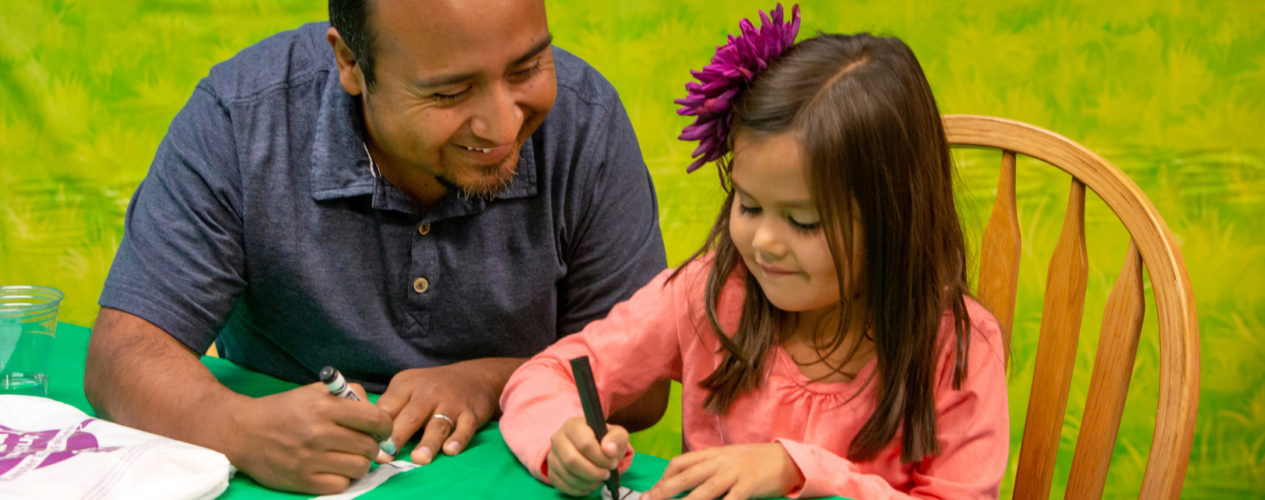 A dad and daughter sit at a table together. The daughter, who is about 7, writes on strips of cloth.