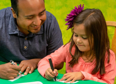 A dad and daughter sit at a table together. The daughter, who is about 7, writes on strips of cloth.