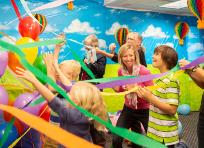 Volunteers and children celebrate among brightly colored streamers and balloons.