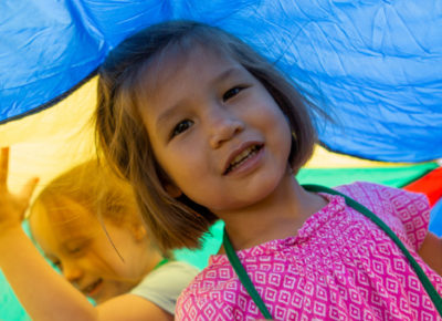 A preschool girl under a colorful parachute at an outreach event.