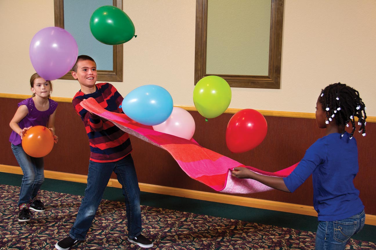 Two children holding a beach towel between them try to keep 6 brightly colored balloons up in the air. One girl is getting ready to add a seventh balloon in the mix.