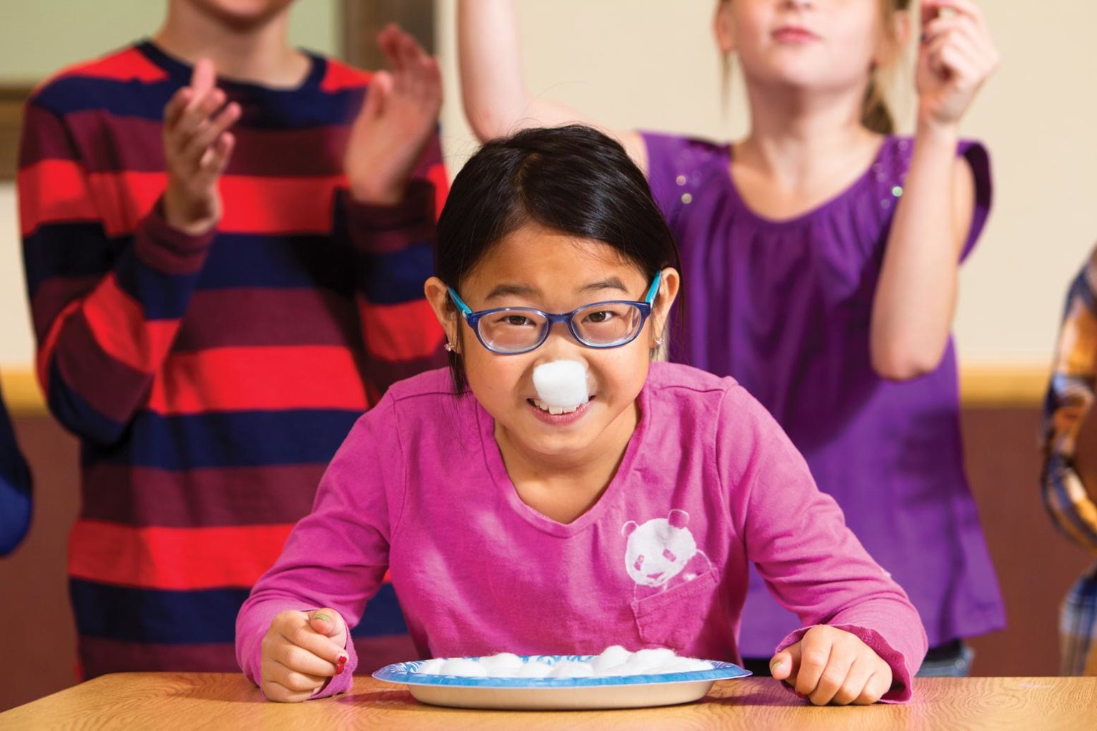 Little girl wearing glasses is smiling as she sits in front of a plate of cotton balls. One cotton ball is stuck to her noise. Her teammates are cheering behind her.