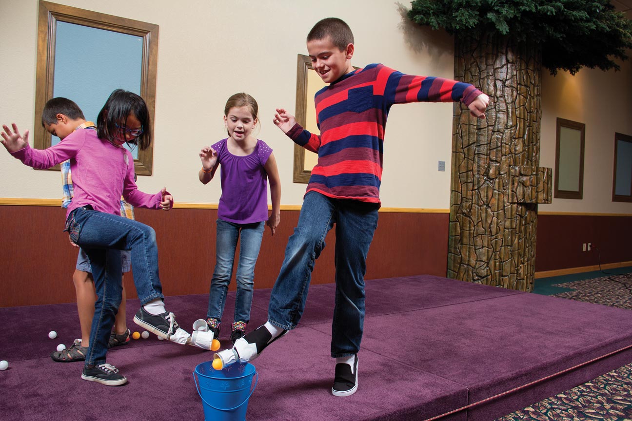 Four kids are trying to scoop up ping-pong balls in the cups on their shoes to place into a bucket.
