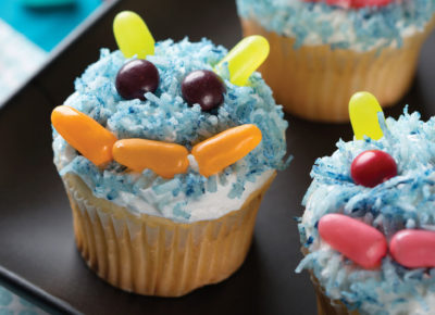 Cupcakes frosted and covered in blue coconut flakes. Candies are used to make a monster mouth, eyes, and ears.