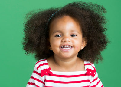 Preschool girl wearing a red and white striped shirt. She's got a huge smile as she sits in front of a green background.