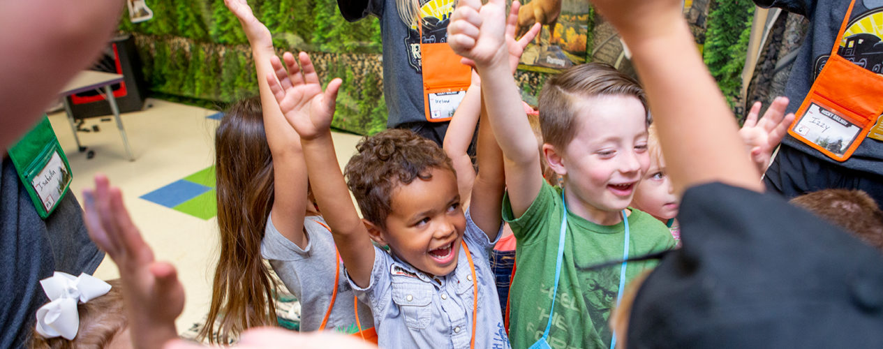 A group of preschoolers with their hands up in celebration.