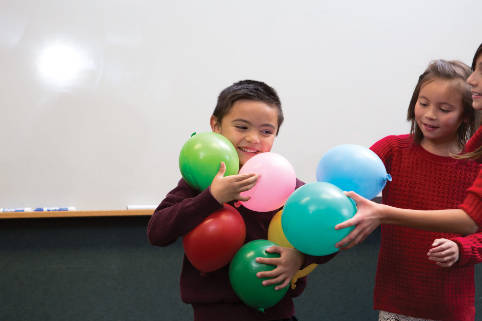 Elementary aged boy trying to hold lots of colorful balloons in his arms. His classmates are trying to add more and more to what he's carrying.