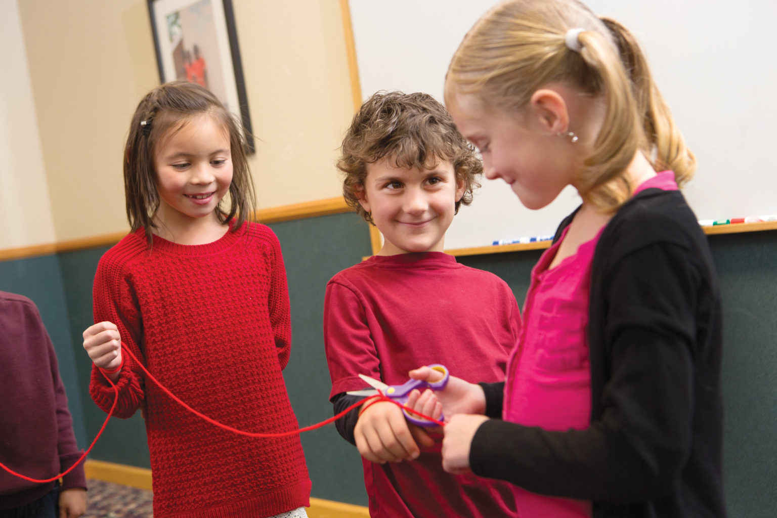 A group of children have yarn tied around their wrists. One girl is freeing them with scissors.