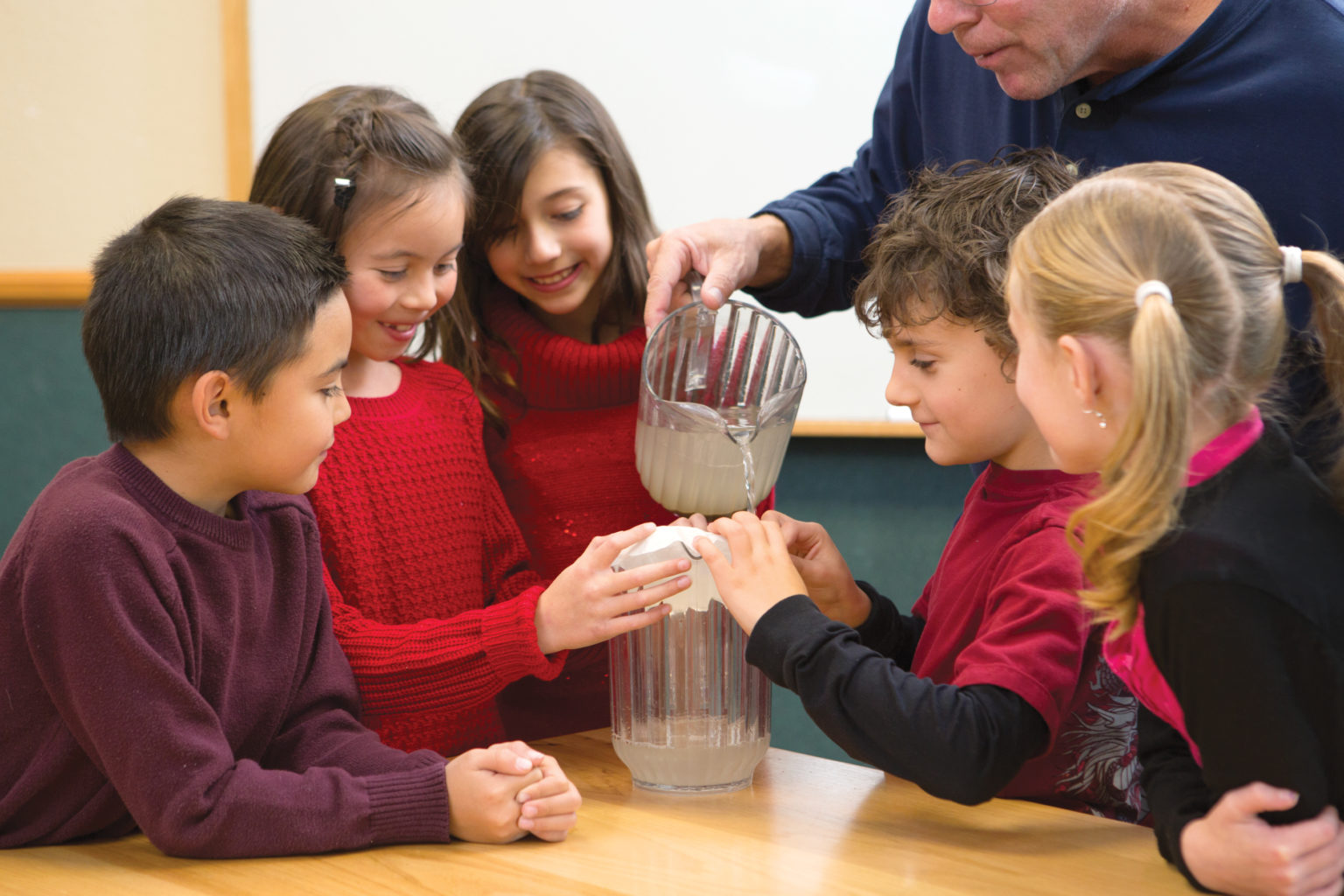A group of elementary aged children gather around as their teacher pours lemonade through a coffee filter.