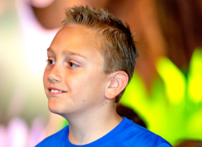 A preteen boy grins. He is standing in front of a brightly colored background.