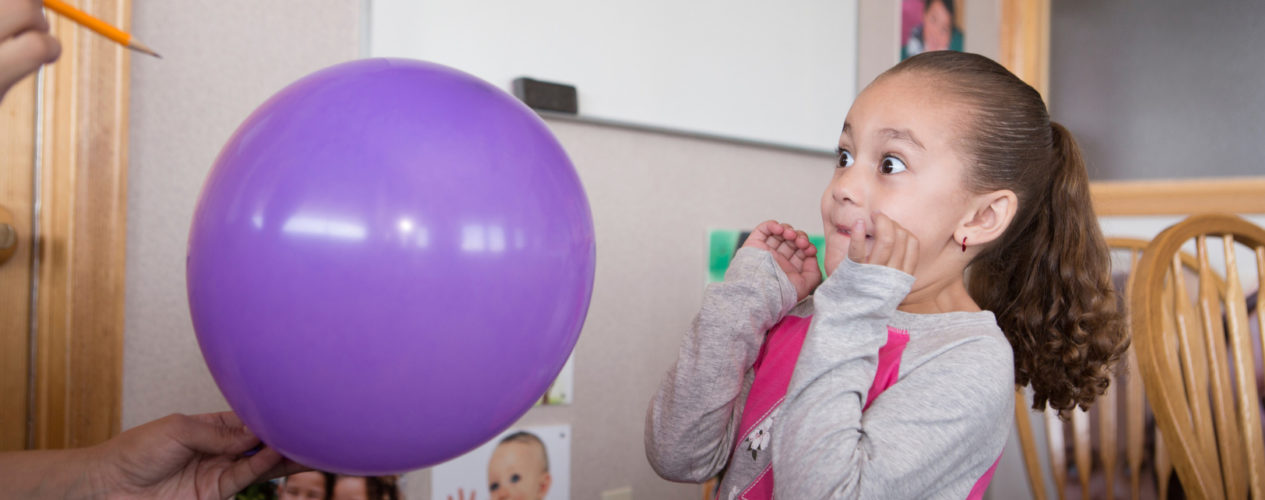 A little girl has a look of fear as a pencil goes to pop a balloon.