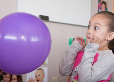 A little girl has a look of fear as a pencil goes to pop a balloon.