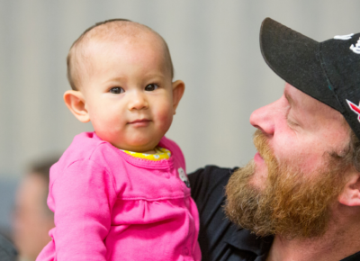 A man with a beard wearing a cap is holding a baby at his church nursery drop off area.