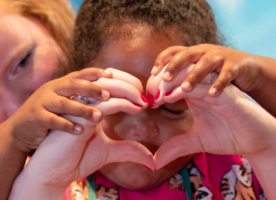 An woman volunteer makes a heart with her hands as a young girl holds the hands to her face and places her hands on top of the volunteer's hands.