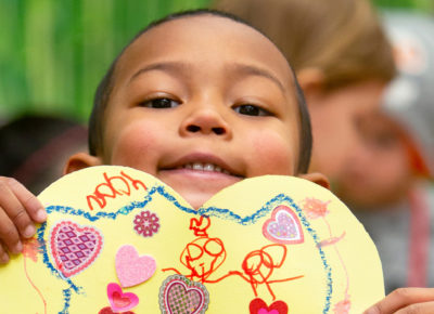 Young elementary boy smiles as he holds a family Valentine's day craft he made.