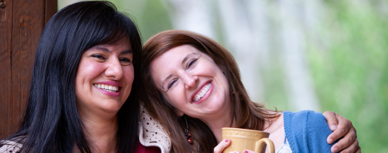One woman with her arm wrapped around another woman's shoulder. The woman receiving the hug is smiling with a mug of coffee in her hands.