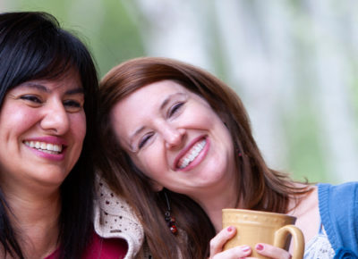 One woman with her arm wrapped around another woman's shoulder. The woman receiving the hug is smiling with a mug of coffee in her hands.