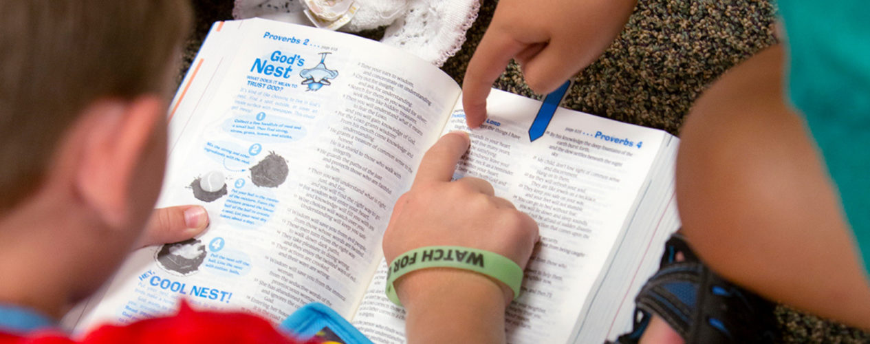 Two elementary boys pointing at a passage in a children's Bible. One boy is wearing a bracelet that says "Watch for God".