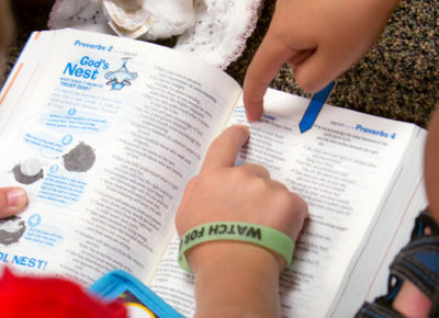 Two elementary boys pointing at a passage in a children's Bible. One boy is wearing a bracelet that says "Watch for God".