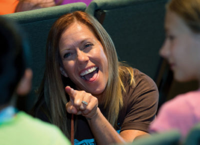 A VBS volunteer smiles as she points at a young boy. He is sitting next to a young girl.