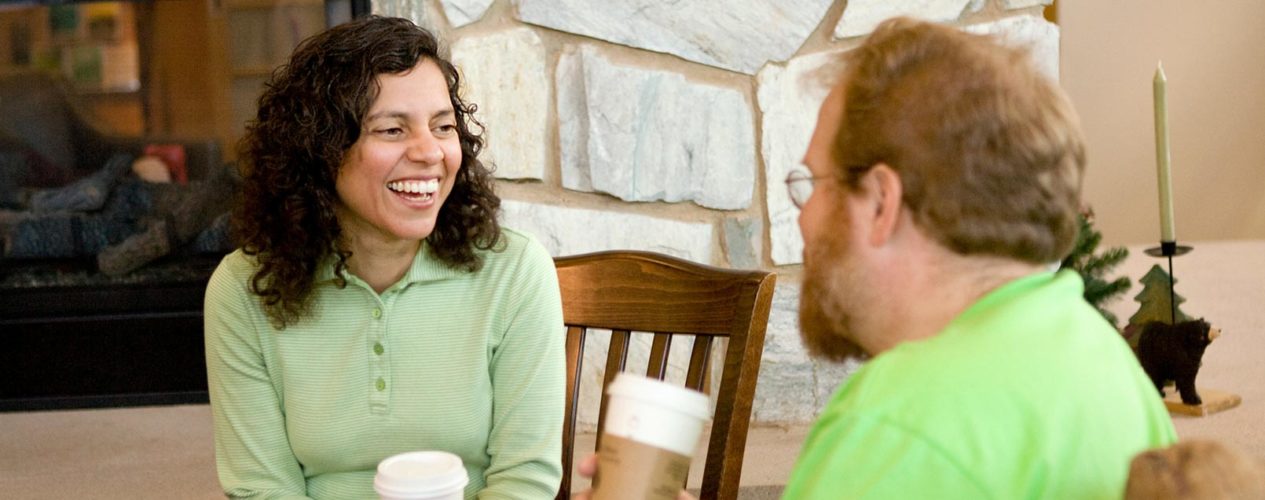Woman talking to a man. They are drinking coffee at a table by the fireplace. She is recruiting him to be a volunteer.