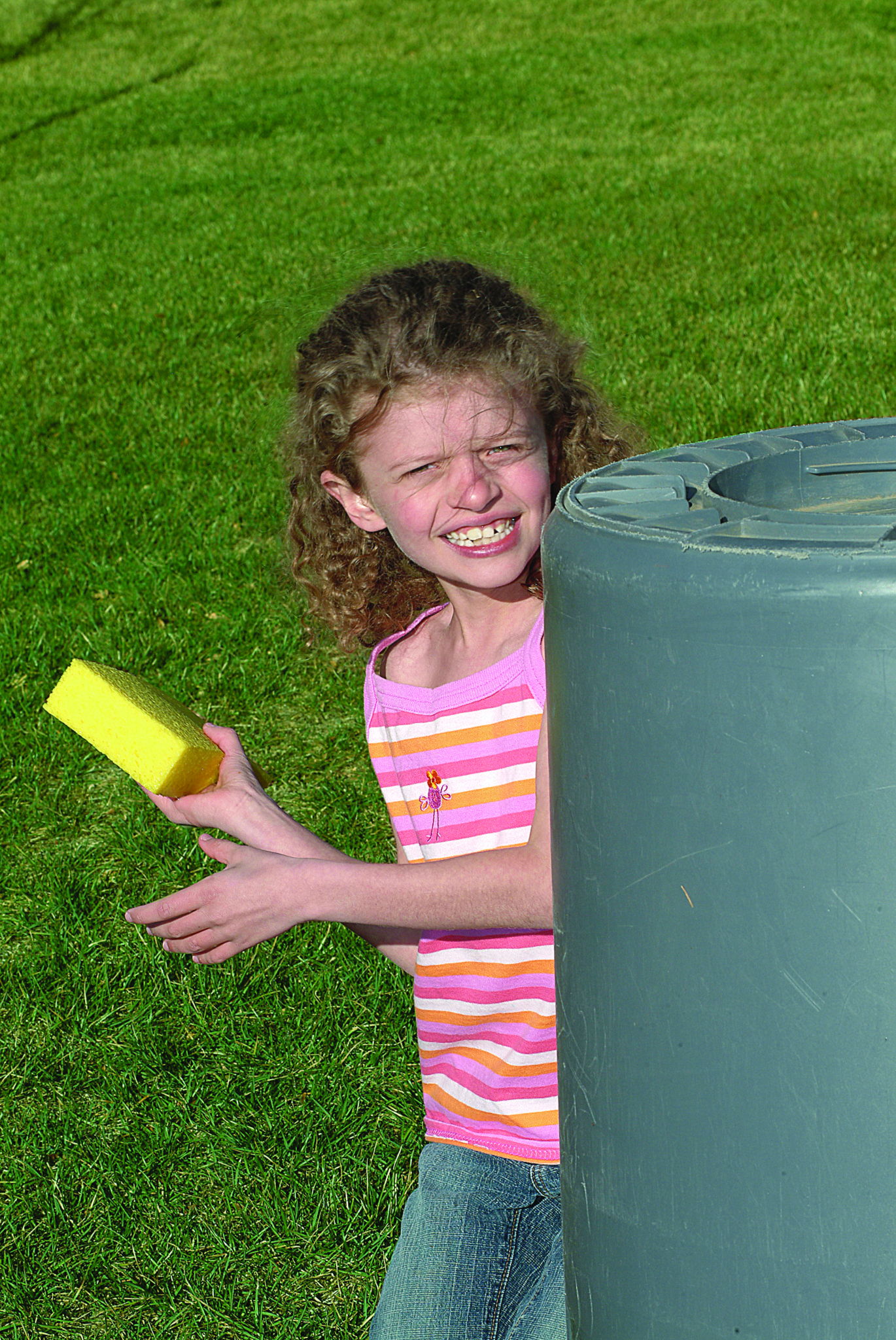 A girl hides behind a huge trash can with a sponge.