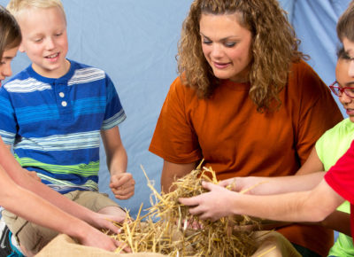 Volunteer sits with four elementary-aged children around a manger. They are feeling the hay in the manger.