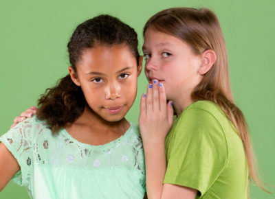 One preteen girl whispers in the ear of another. They are staring directly at the camera.