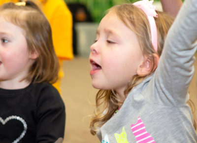 Two preschool girls sing. One has her arm stretched up in joy.