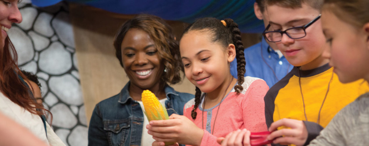 Preteen girl holding a plastic ear of corn as her peers grab other plastic food. Their teachers look on smiling.